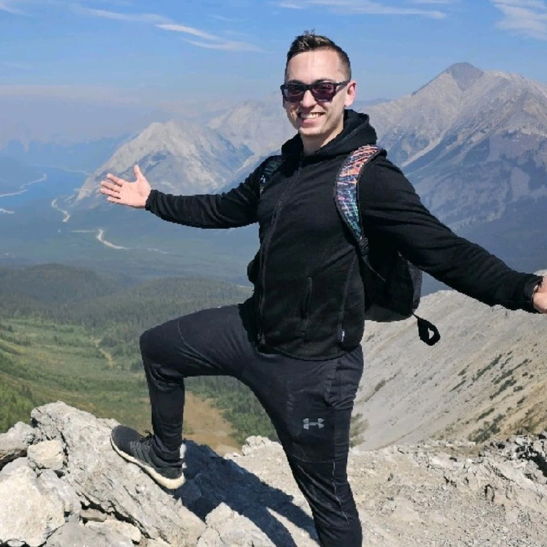 David Minin on a mountain summit with arms open, sunglasses, and a backpack against a clear sky